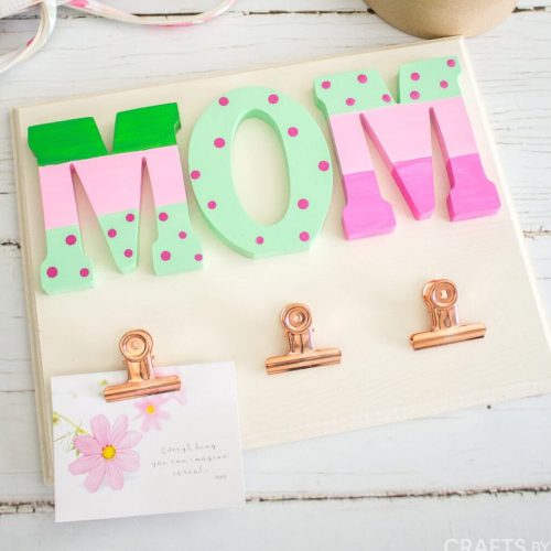 A craft project spelling "mom" in large, colorful wooden letters on a beige board, with decorative clips and a flower card on a wooden table.