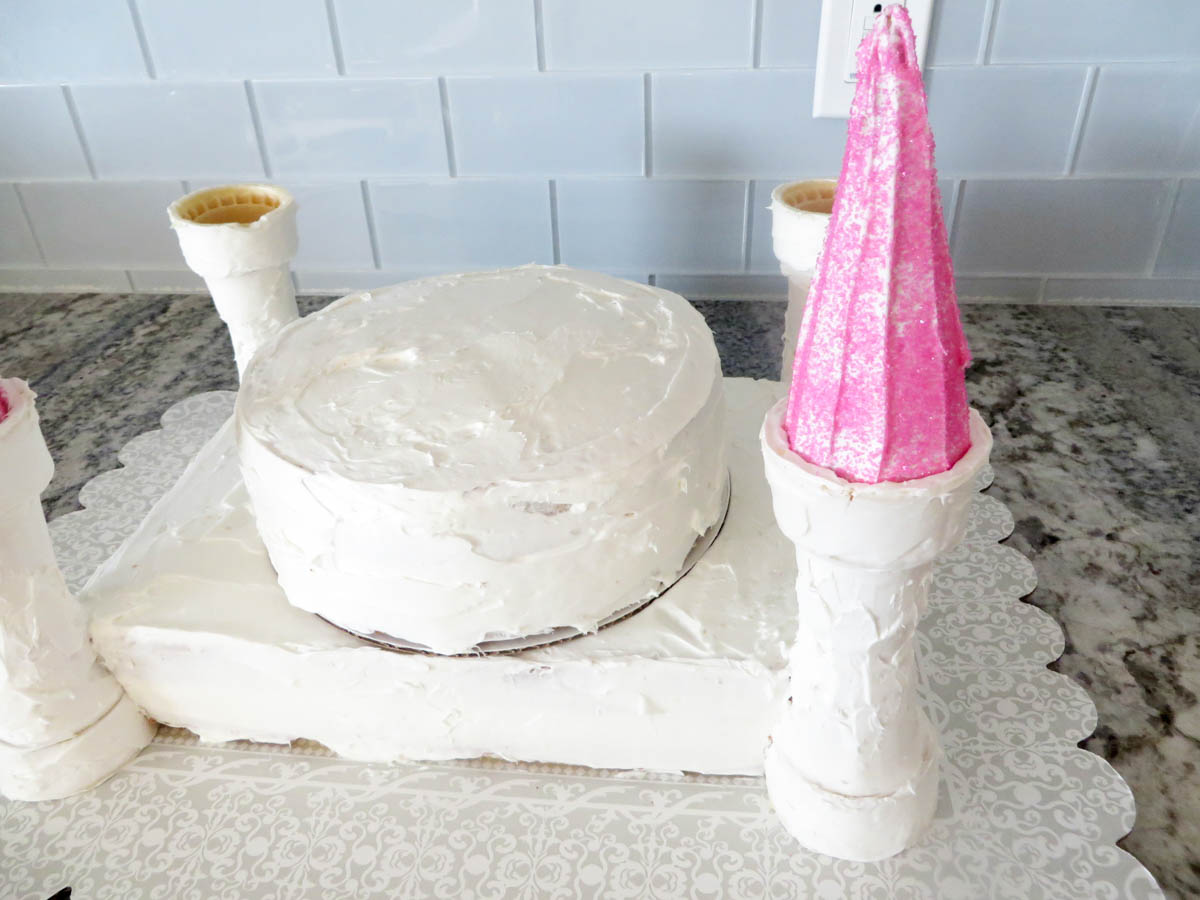 A homemade cake shaped like a castle, featuring a round body and three cone-topped towers, decorated with white frosting and a pink glittery cone.