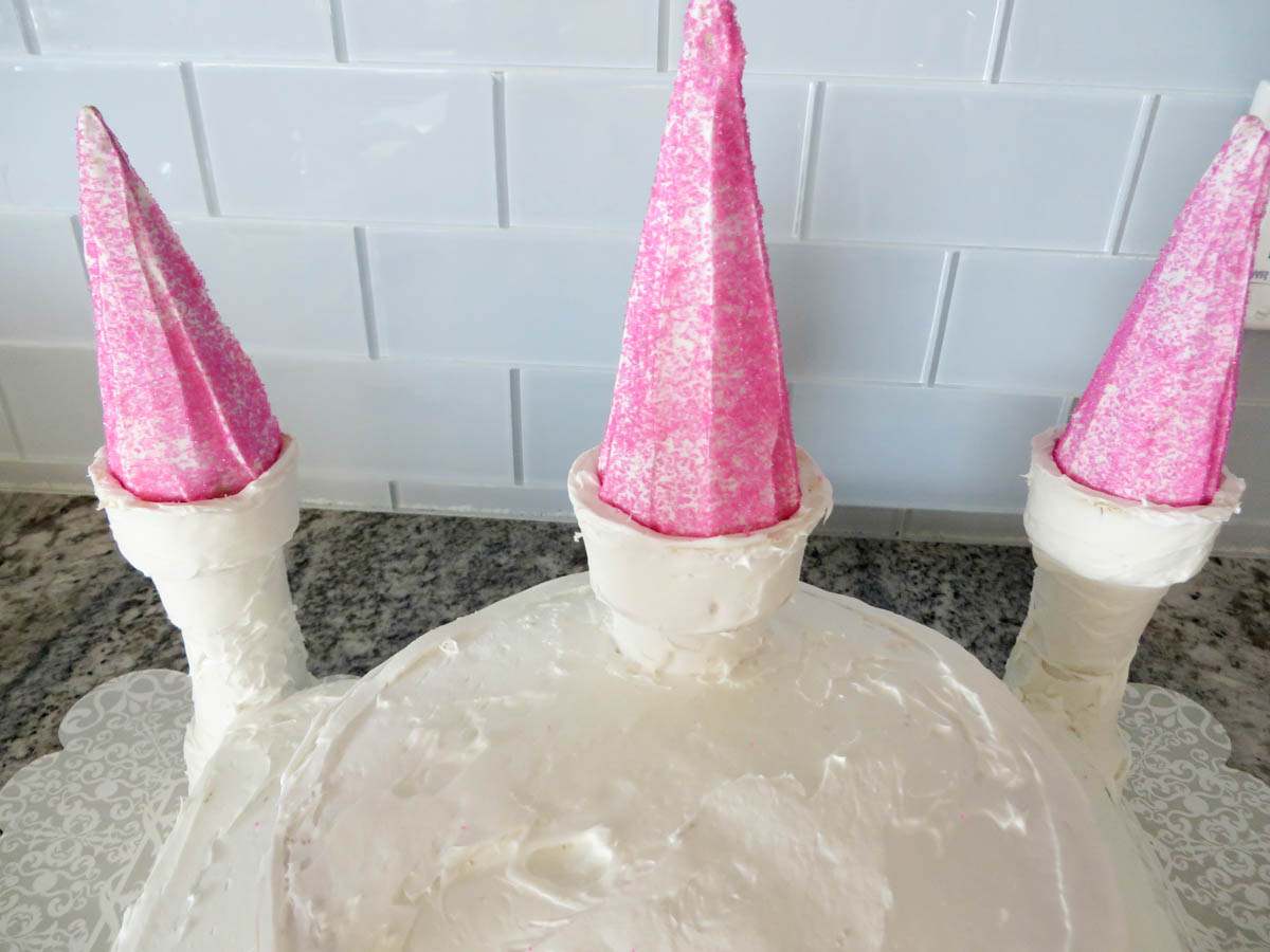 A cake decorated with white frosting and topped with three pink ice cream cones to resemble a castle's turrets.