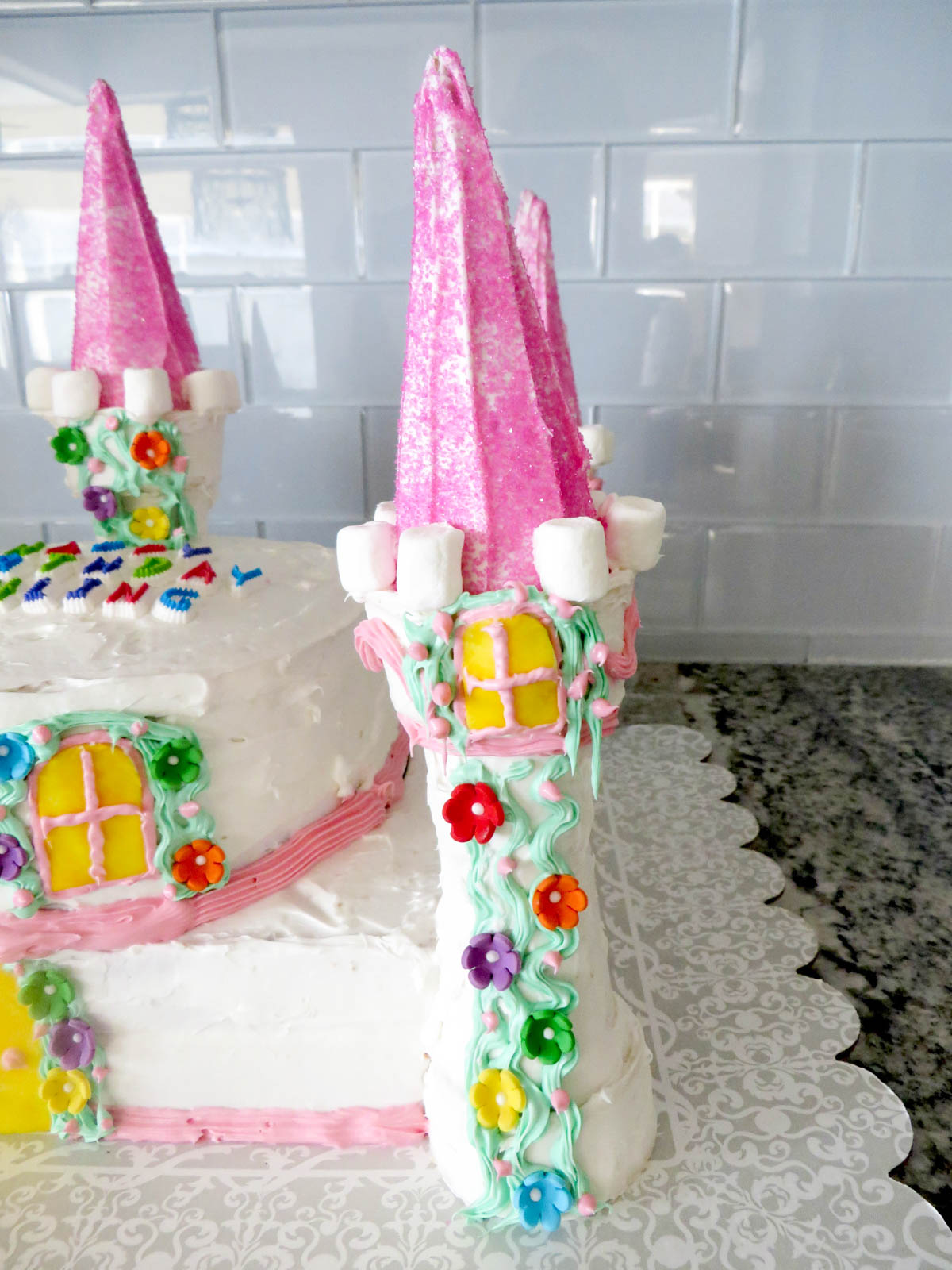 A whimsical castle-shaped cake with pink turrets, colorful flowers, and marshmallow details on a kitchen counter.