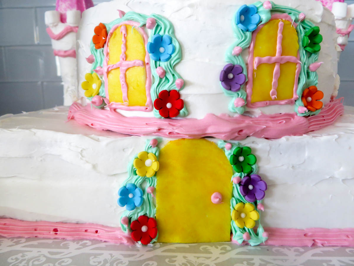 Two-tier cake decorated to resemble a house with white frosting, colorful piped windows and doors, and multicolored flowers.