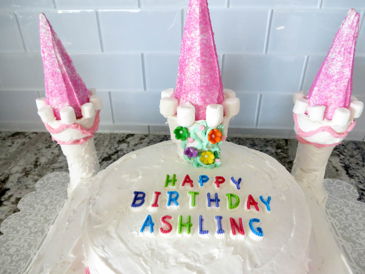 Birthday cake decorated with "happy birthday ashling" in colorful letters, topped with three pink ice cream cone towers and multi-colored flowers on a kitchen counter.