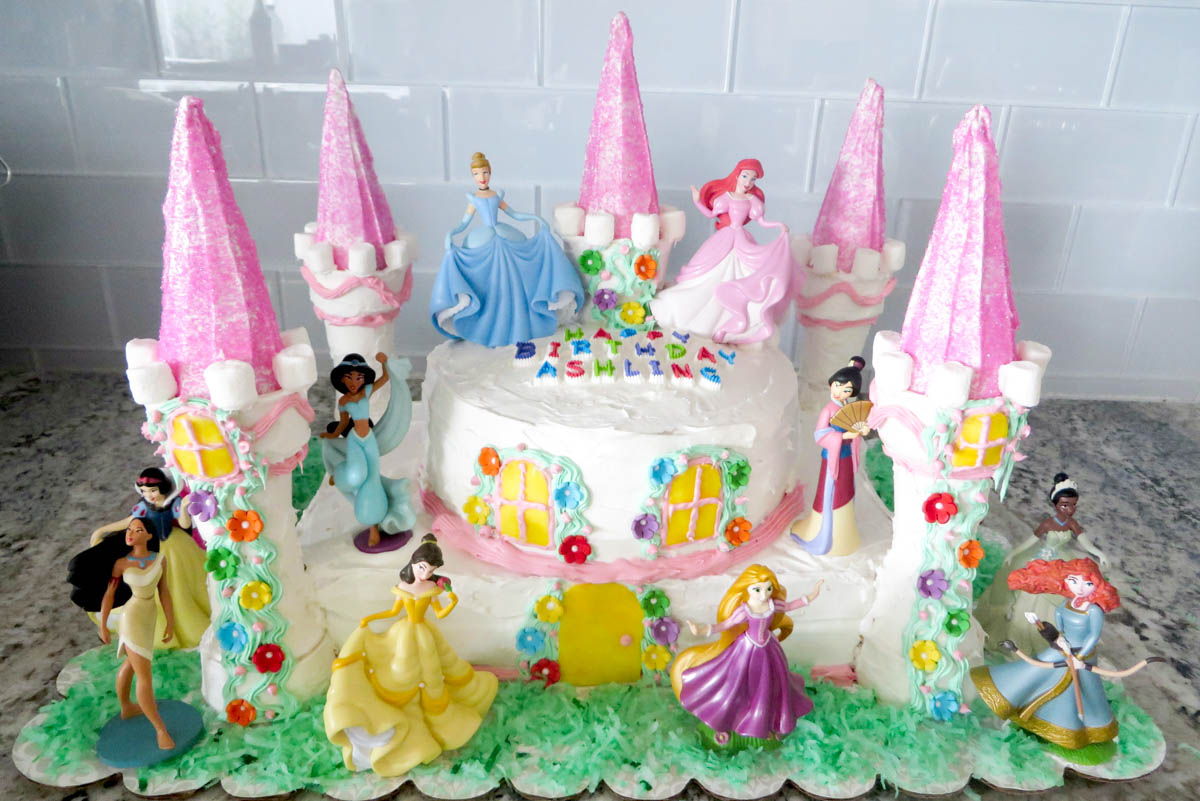 A colorful birthday cake decorated with figures of various disney princesses and tall pink towers, personalized with the name "ashlyn.