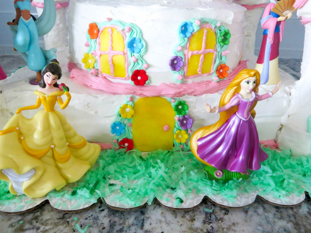 A colorful birthday cake decorated with figures of princesses belle and rapunzel, surrounded by vibrant flowers and green coconut shreds resembling grass.