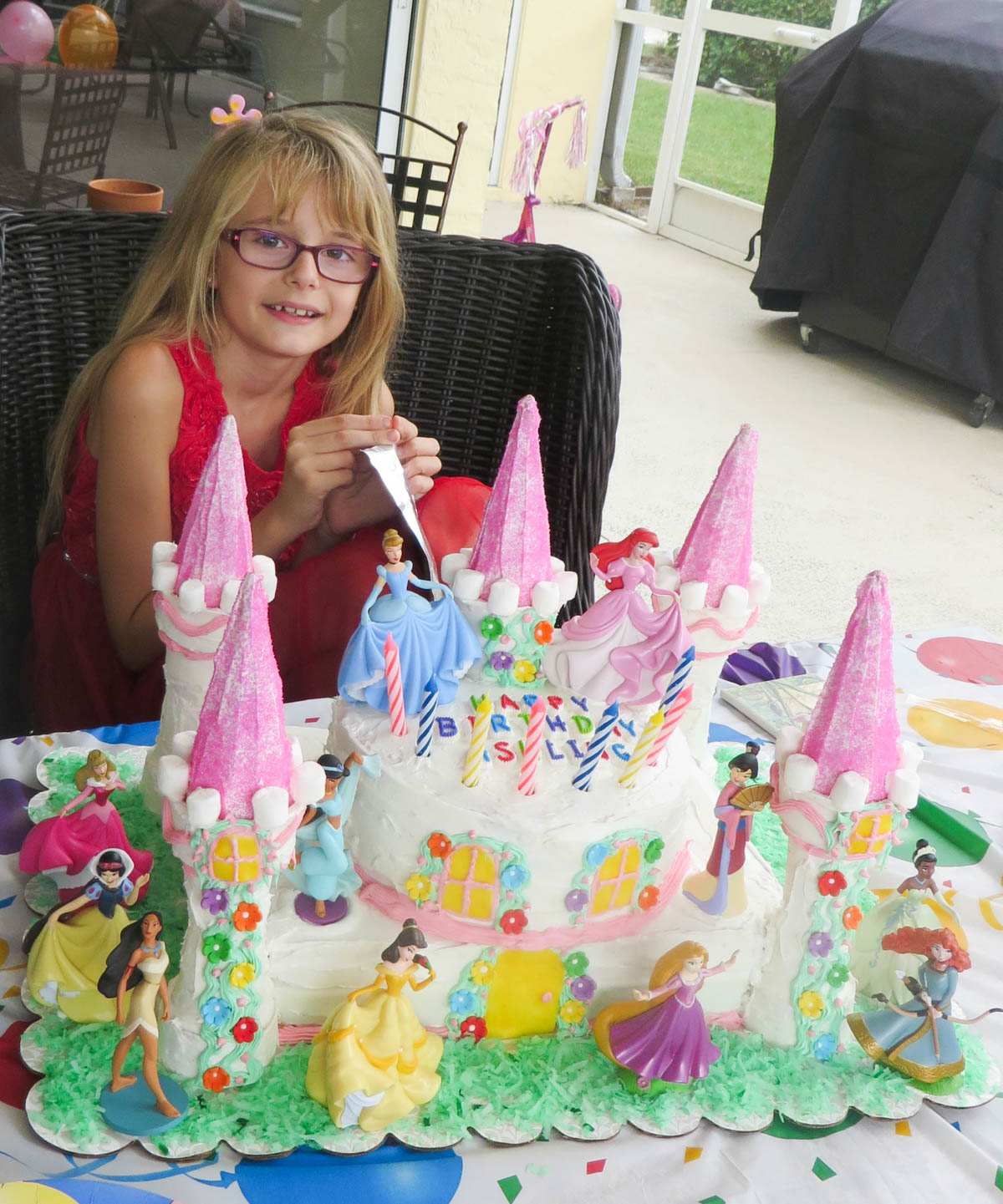 Young girl in red, wearing glasses, sits before a birthday cake adorned with princess figurines and colorful decorations, smiling while holding a candle.