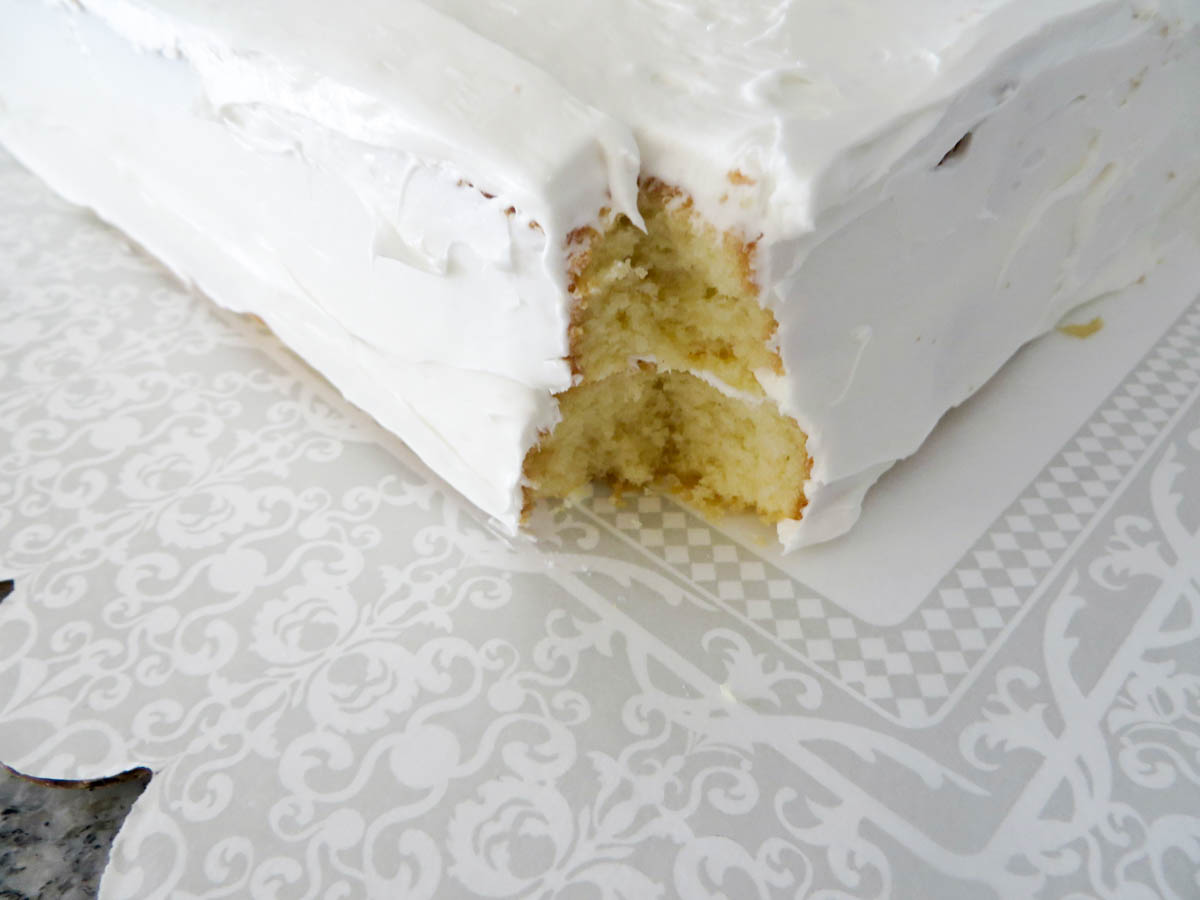 A white frosted cake with a missing piece, showing its inner fluffy yellow layers, on a patterned grey and white surface.