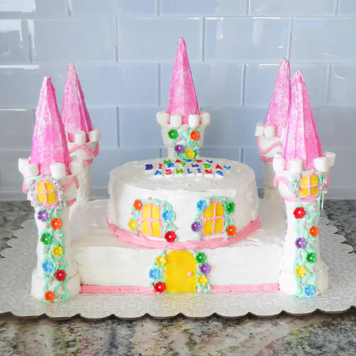 A colorful castle-themed birthday cake with pink towers and decorative candy elements on a kitchen counter.