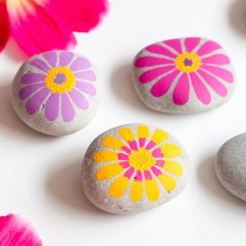 Three painted stones with floral designs in pink and yellow, accompanied by pink flower petals, on a white background.