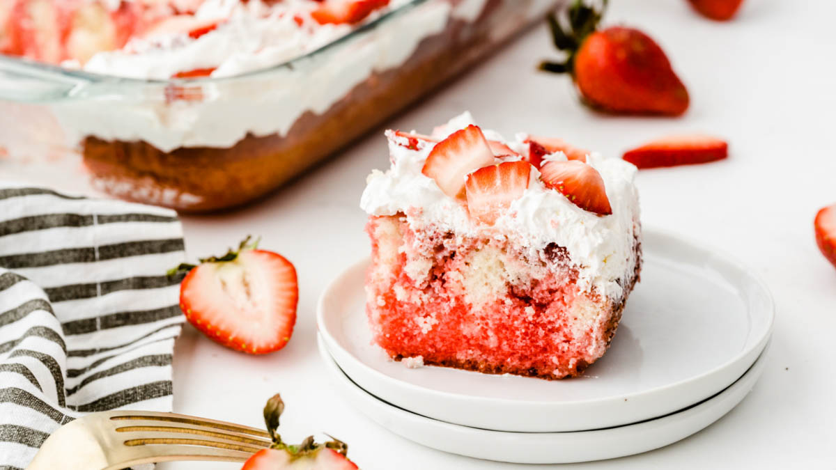 A slice of strawberry poke cake with whipped cream and fresh strawberries on a white plate, beside whole strawberries and a cake pan.