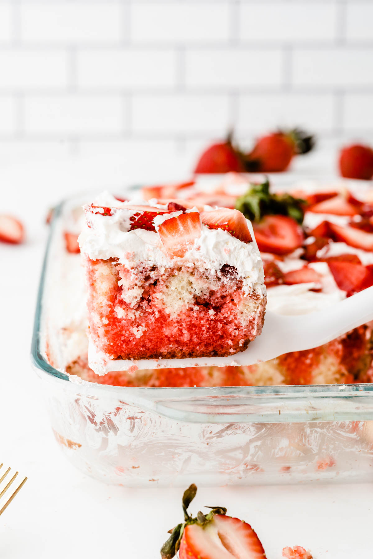 A strawberry poke cake with a slice being lifted, revealing its layers and fresh strawberry topping.
