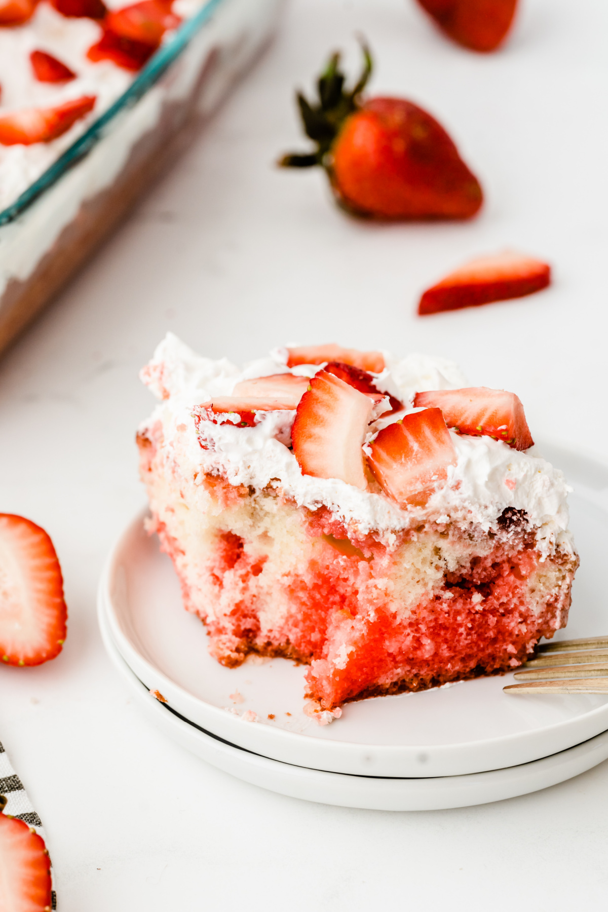 A slice of strawberry poke cake with whipped cream and fresh strawberry slices on a white plate.