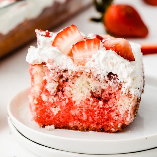 A slice of strawberry cake topped with whipped cream and fresh strawberry slices on a white plate.