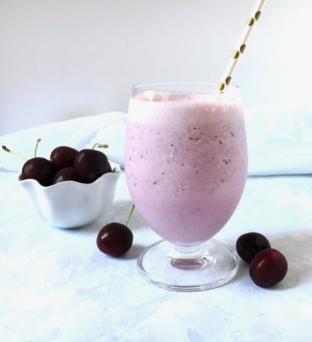 A glass of cherry smoothie with a straw, accompanied by a bowl of fresh cherries on a white surface.
