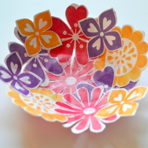 A colorful bowl filled with decorative paper flowers in shades of orange, purple, red, and pink, arranged artistically to form a vibrant display.