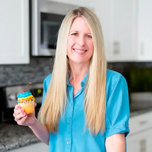 A woman with long blonde hair, wearing a blue shirt, smiling and holding a decorated cupcake in a modern kitchen.