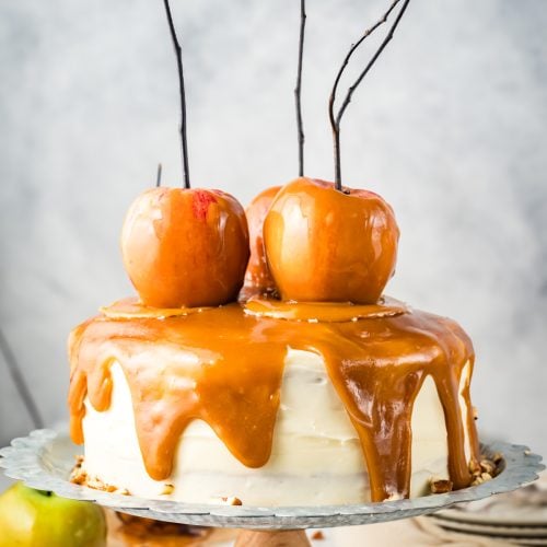 A frosted cake topped with caramel drizzle and three caramel-dipped apples on sticks sits on a decorative metal plate.