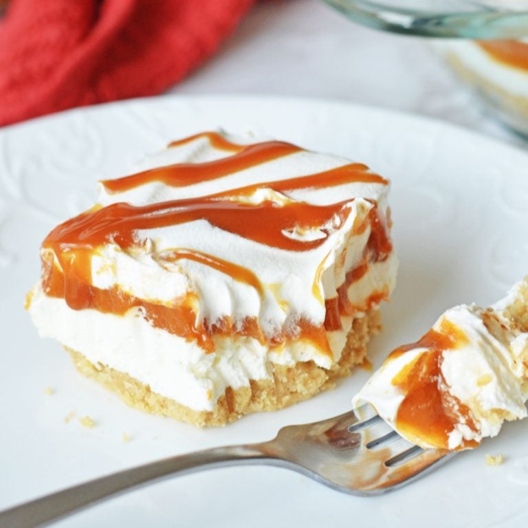 A plated dessert features a square slice of layered cream and caramel on a crumbly base with a fork holding a small piece nearby.