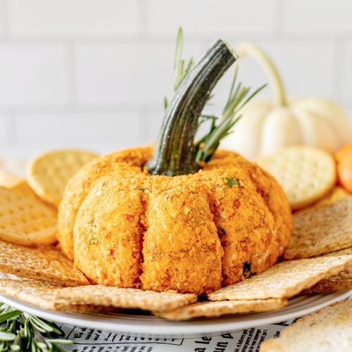 A pumpkin-shaped cheese ball with a stem garnish is surrounded by an assortment of crackers on a platter.