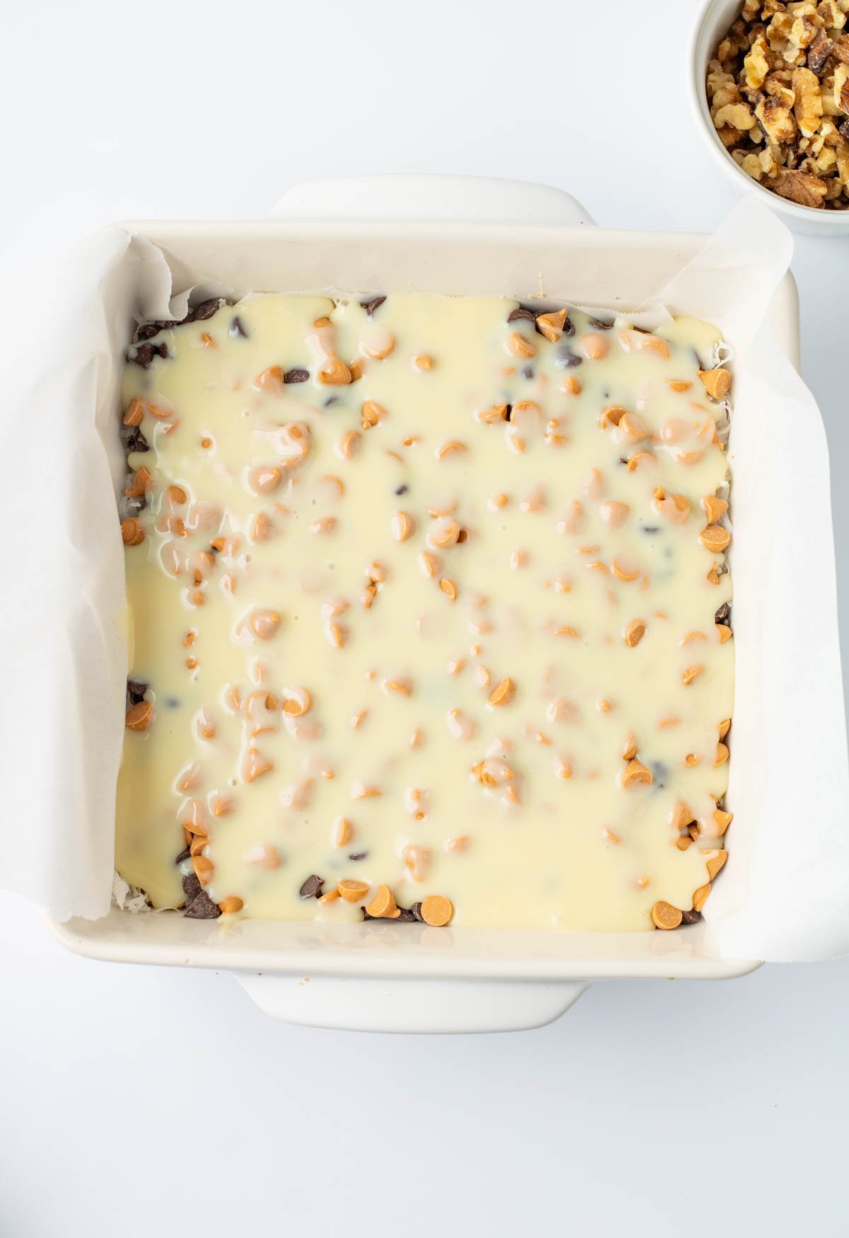A square baking dish lined with parchment paper contains a dessert topped with melted white chocolate and butterscotch chips. A bowl of chopped nuts is visible in the corner.