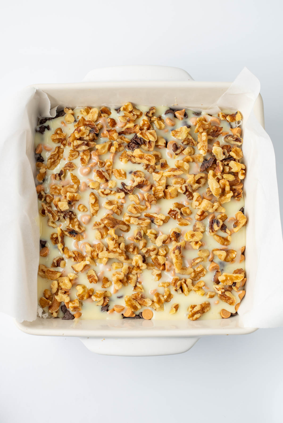 A square baking dish lined with parchment paper, filled with a dessert topped with chopped walnuts and a layer of melted white chocolate.