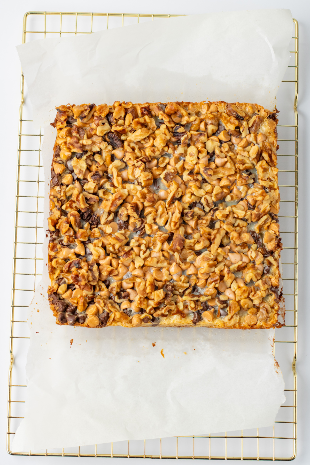 A square nut-topped dessert bar sits on parchment paper atop a cooling rack.