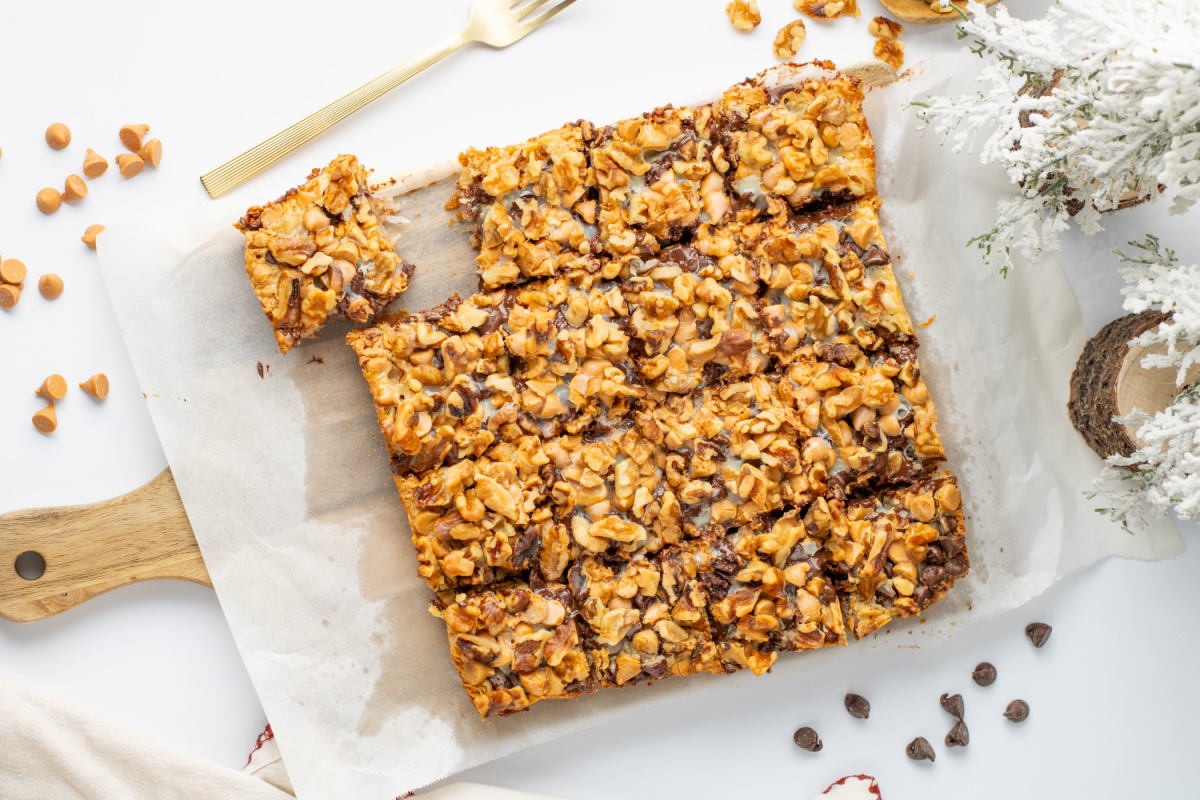 A tray of nut-topped dessert bars cut into squares sits on parchment paper, with a gold fork and scattered chocolate and butterscotch chips nearby.