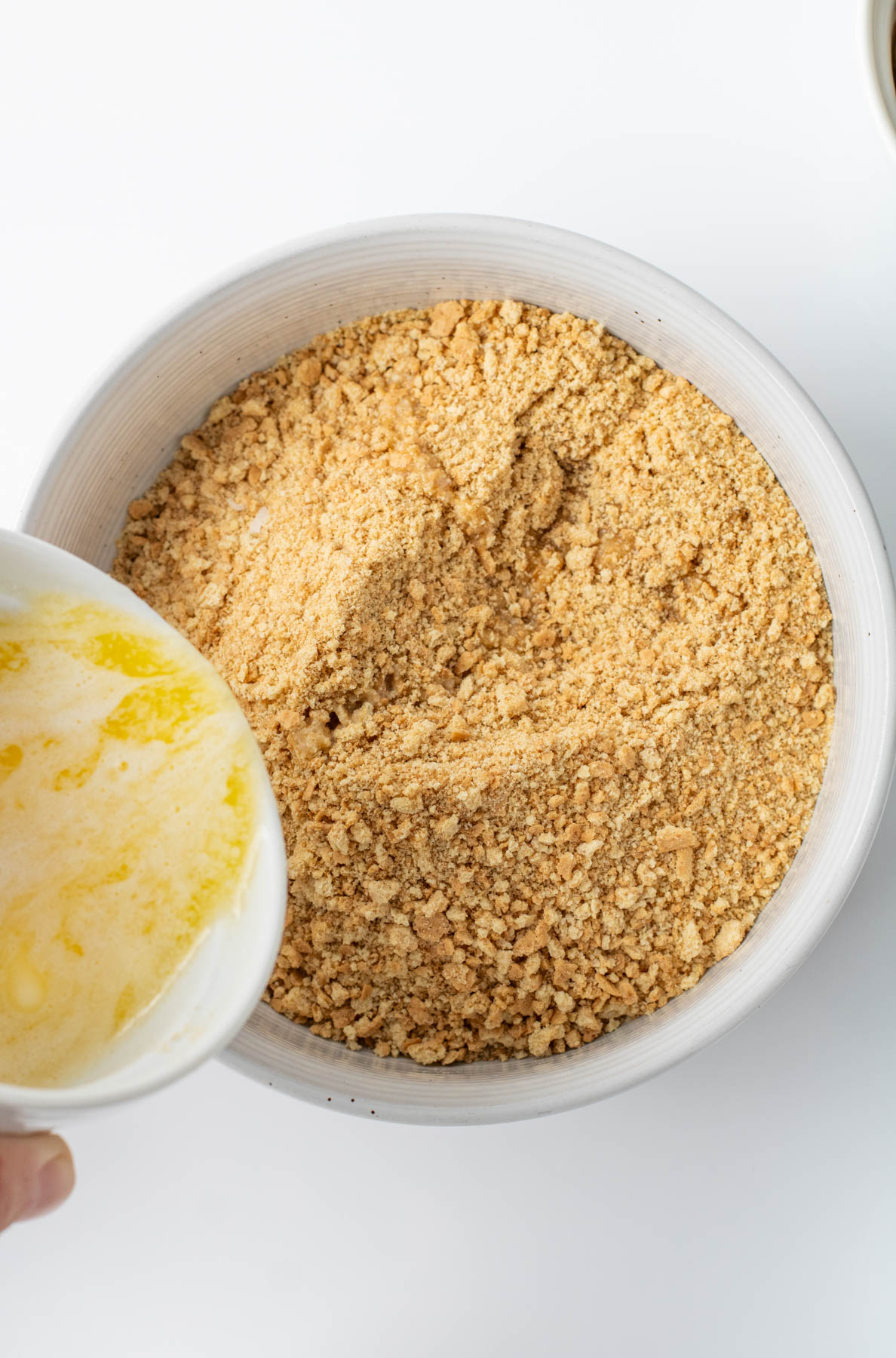 Crushed graham crackers in a white bowl with melted butter being poured in from a separate bowl.