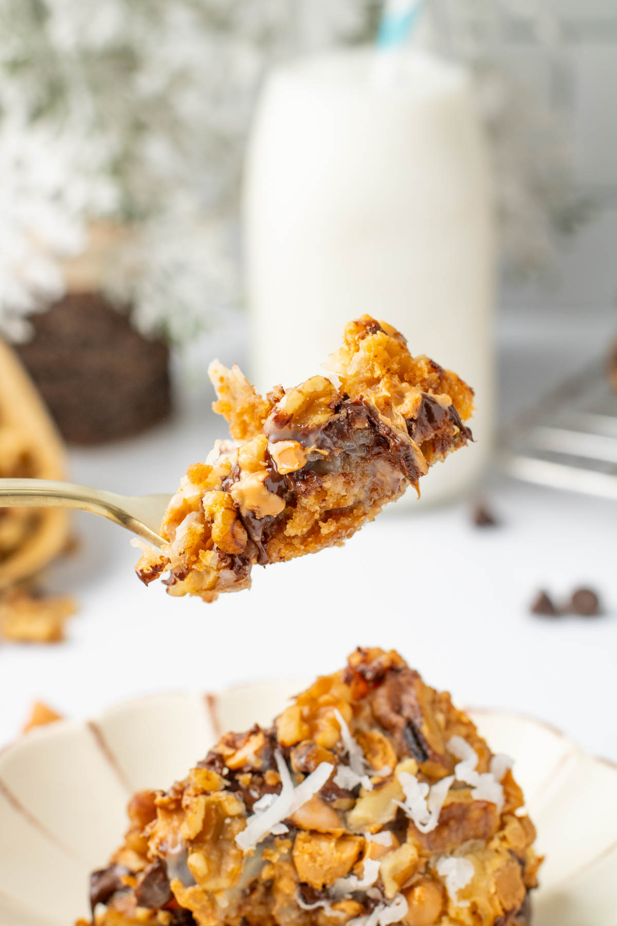 A fork holds a piece of a dessert bar with chocolate chips and coconut flakes; a glass bottle of milk is in the blurred background.