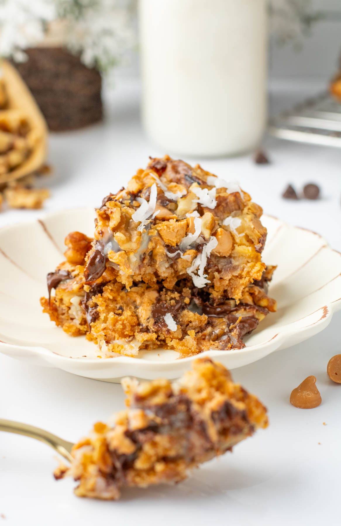 A plate with two stacked dessert bars topped with shredded coconut, chocolate chips, and butterscotch chips; a fork holds a bite in the foreground.