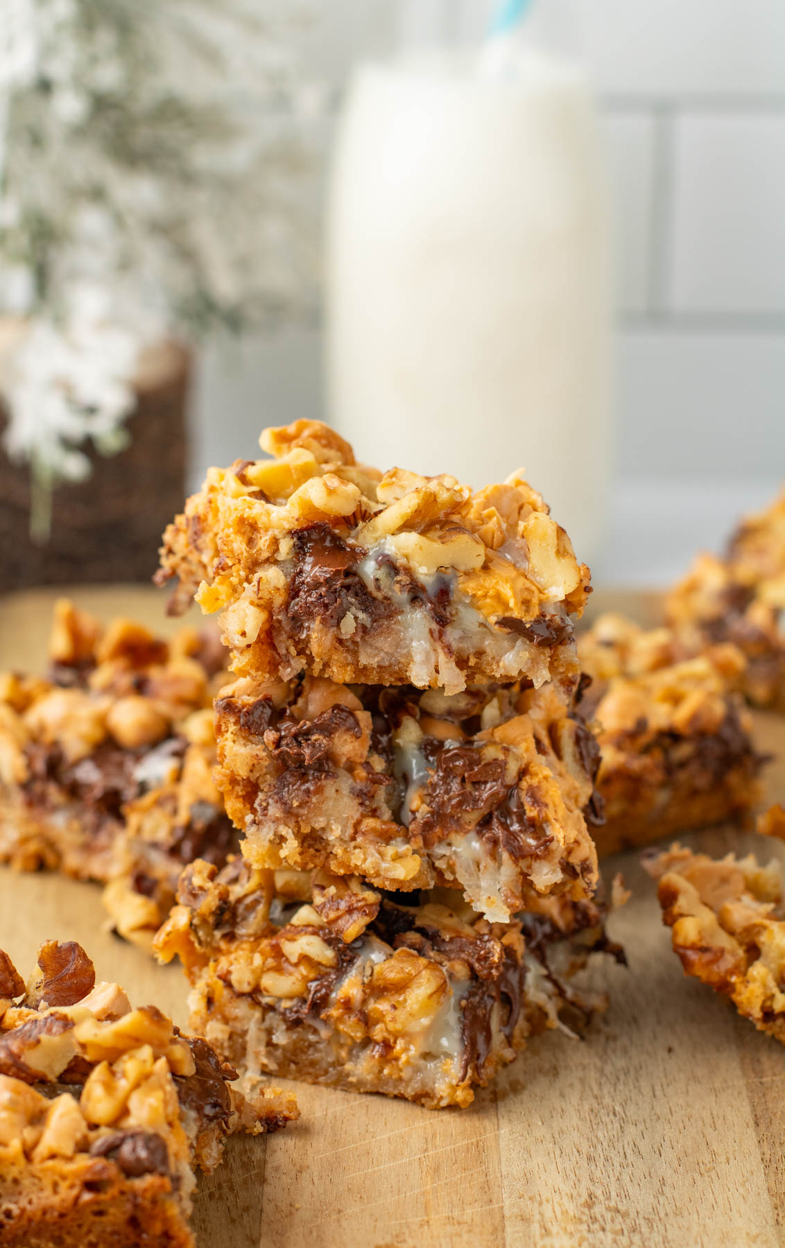 Three dessert bars with a crumbly texture, chocolate chips, nuts, and a gooey filling are stacked on a wooden surface. A glass of milk is in the background.