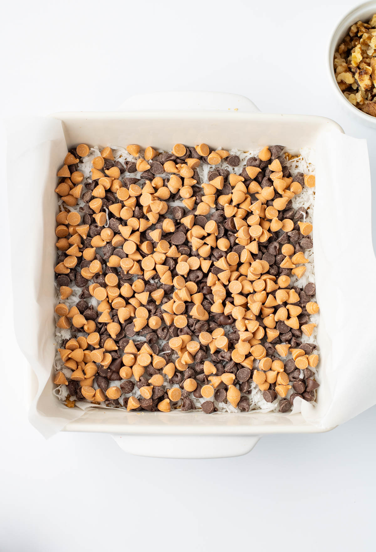 A square baking dish lined with parchment paper, filled with a mixture topped with chocolate chips and butterscotch chips. A small bowl of chopped walnuts is nearby.