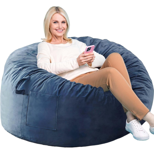 A woman sits on a large, blue bean bag chair, holding a smartphone and smiling. She wears a light sweater, tan pants, and white sneakers.
