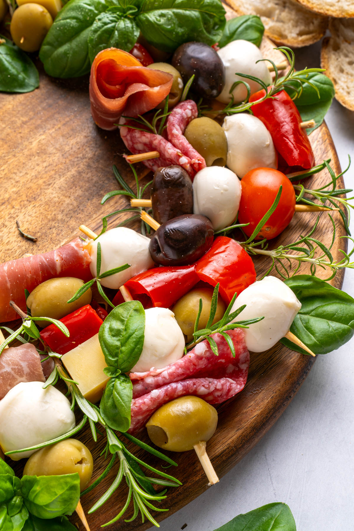 A wooden board displays skewers with mozzarella, olives, salami, tomatoes, and basil, arranged in a wreath shape, with pieces of bread nearby.