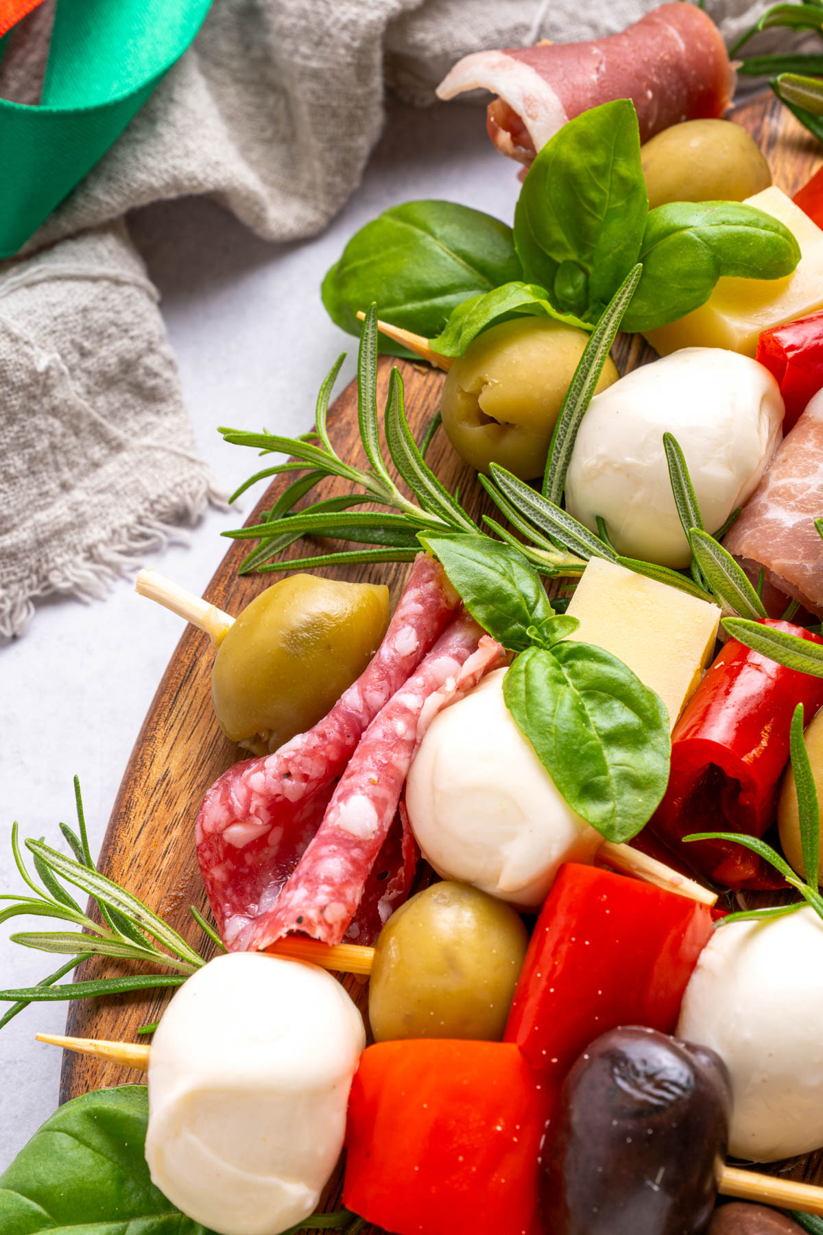 Close-up of an antipasto platter with mozzarella balls, olives, salami, cheese, prosciutto, roasted red peppers, fresh basil, and rosemary on a wooden board.