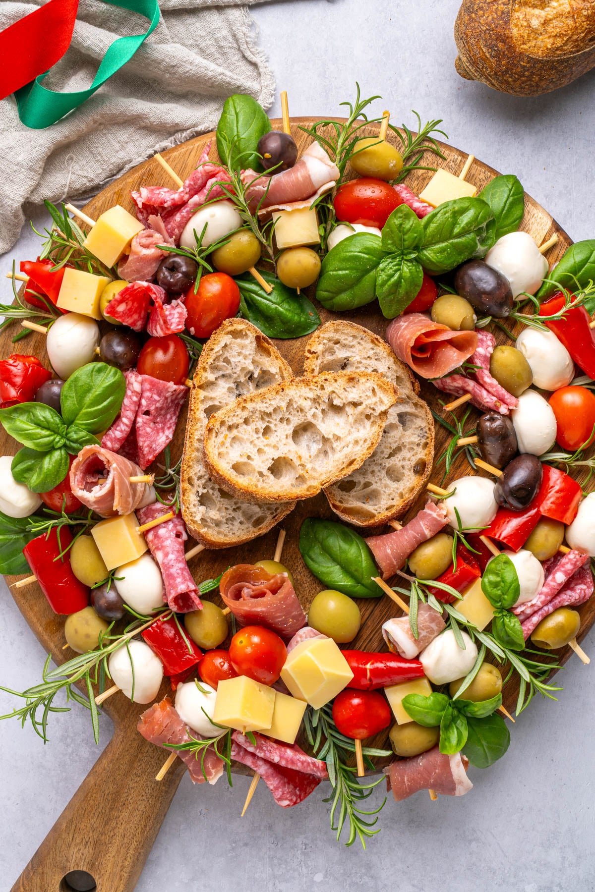 A round wooden board with skewers of mozzarella, tomatoes, olives, cheese, salami, and basil, arranged around two slices of rustic bread in the center.