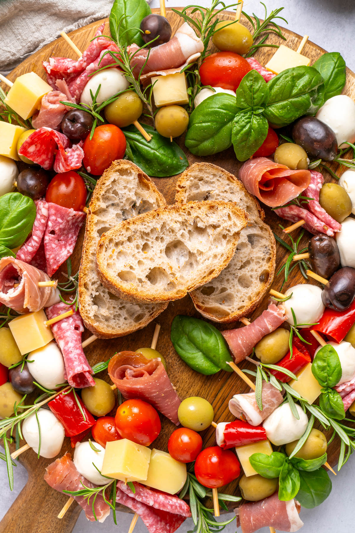A wooden board topped with skewers of cheese, meats, tomatoes, olives, basil, and mozzarella, arranged in a ring around three slices of rustic bread.