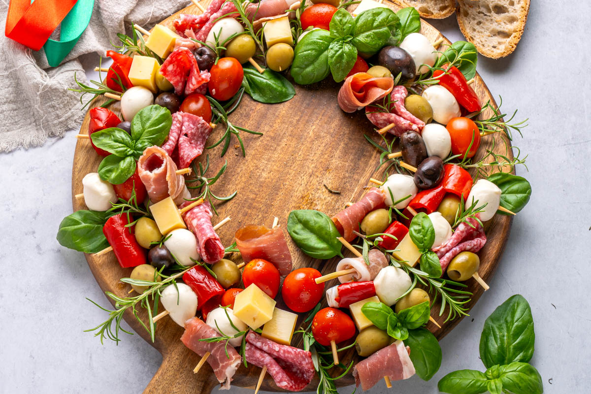 Charcuterie wreath with skewers of cured meats, cheese, tomatoes, olives, and basil arranged on a wooden board, with bread slices on the side.