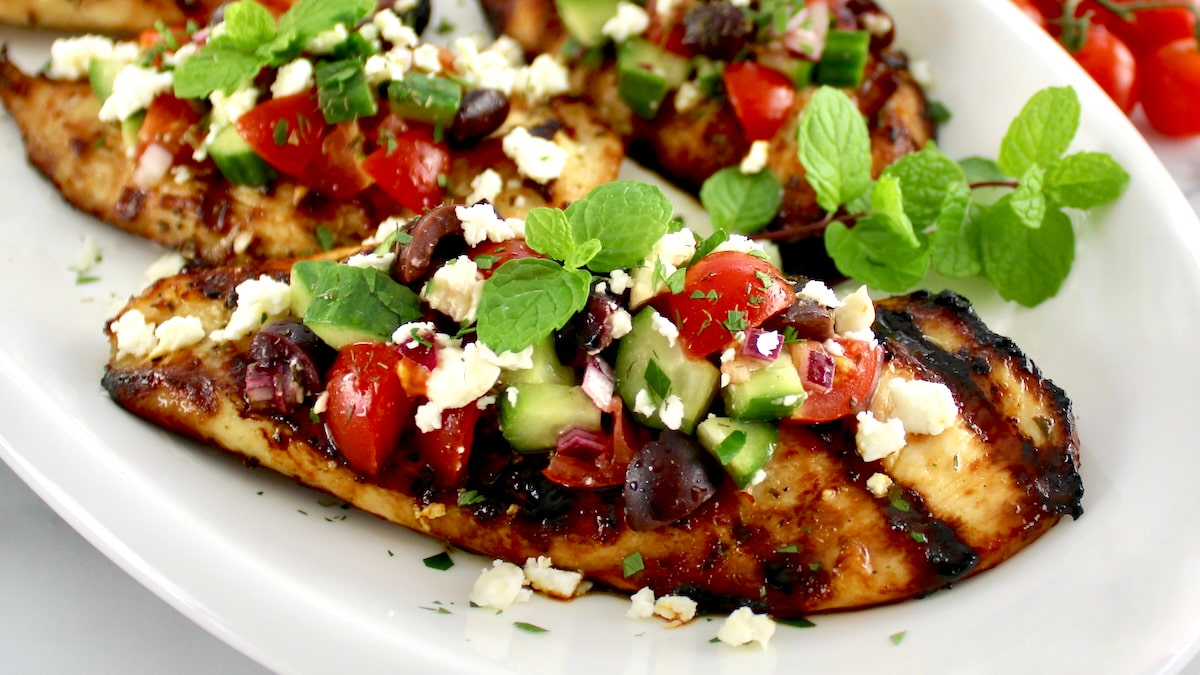 Grilled chicken breasts topped with a mixture of chopped tomatoes, cucumbers, olives, feta cheese, red onions, and fresh mint on a white plate.
