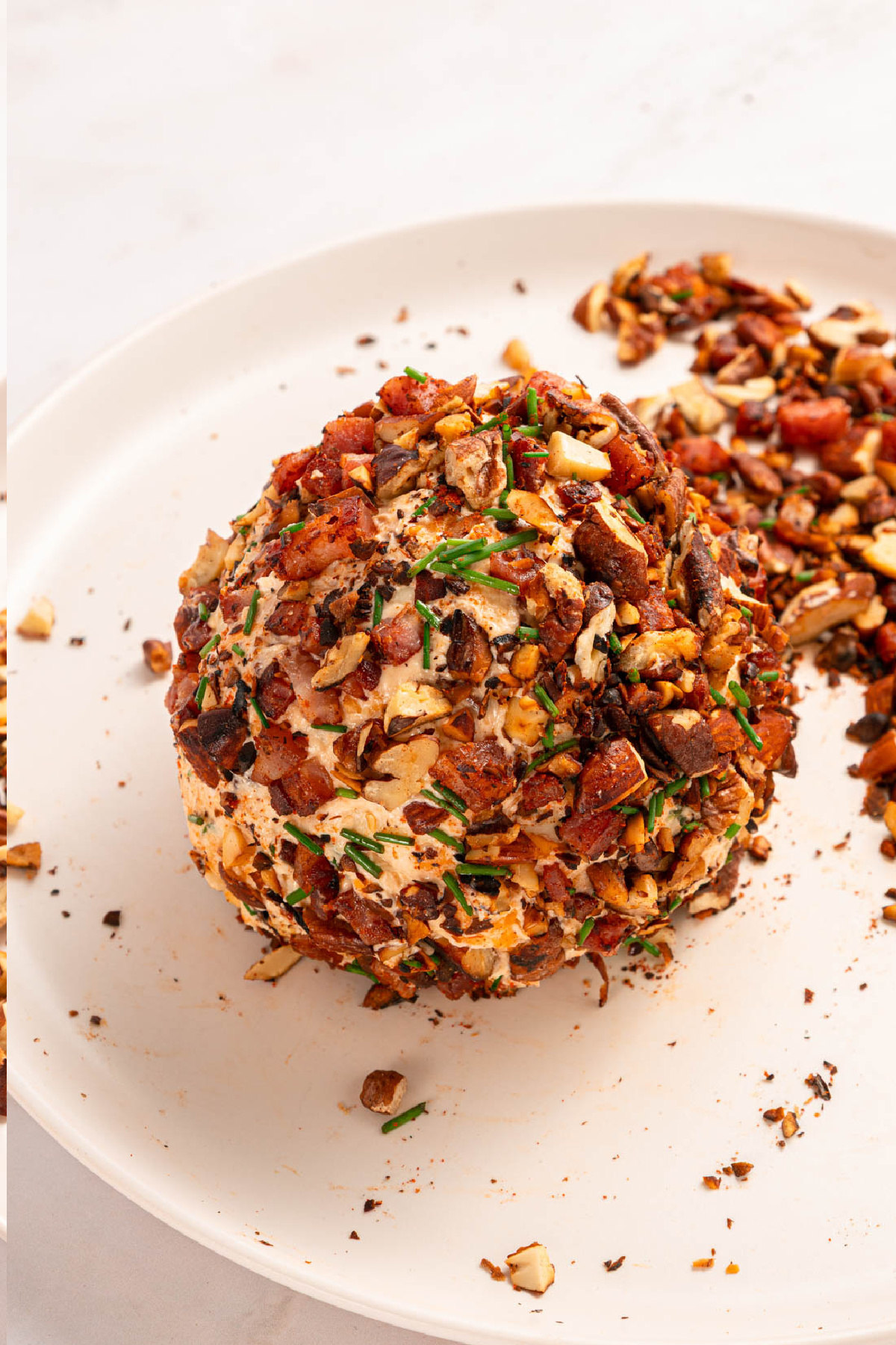 A round cheese ball coated with chopped nuts, bacon, and herbs sits on a white plate, surrounded by scattered pieces of the coating.