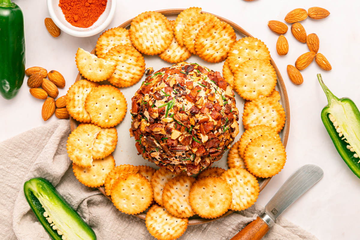 A cheese ball coated with chopped nuts and herbs, surrounded by round crackers, almonds, and halved jalape&ntilde;os on a wooden platter.