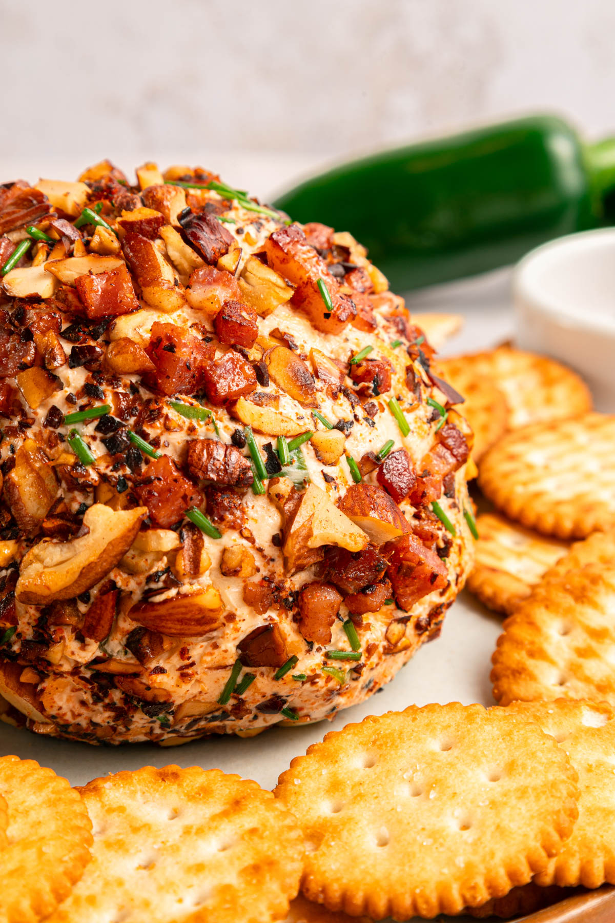 A cheese ball coated with chopped nuts, bacon, and herbs is served on a plate with round crackers and a green jalape&ntilde;o in the background.