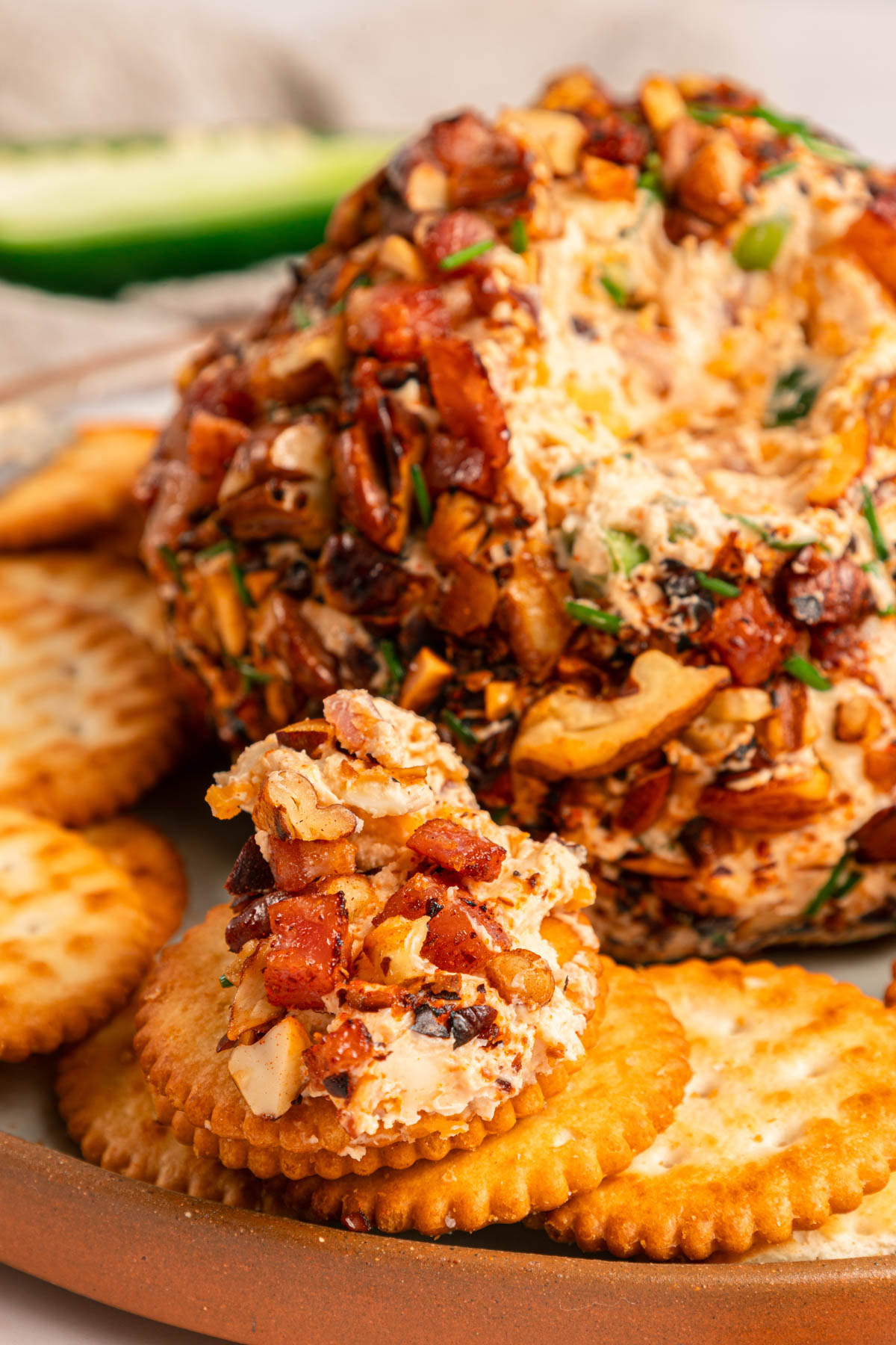 A cheese ball covered in chopped nuts and herbs is served with round crackers on a brown plate.