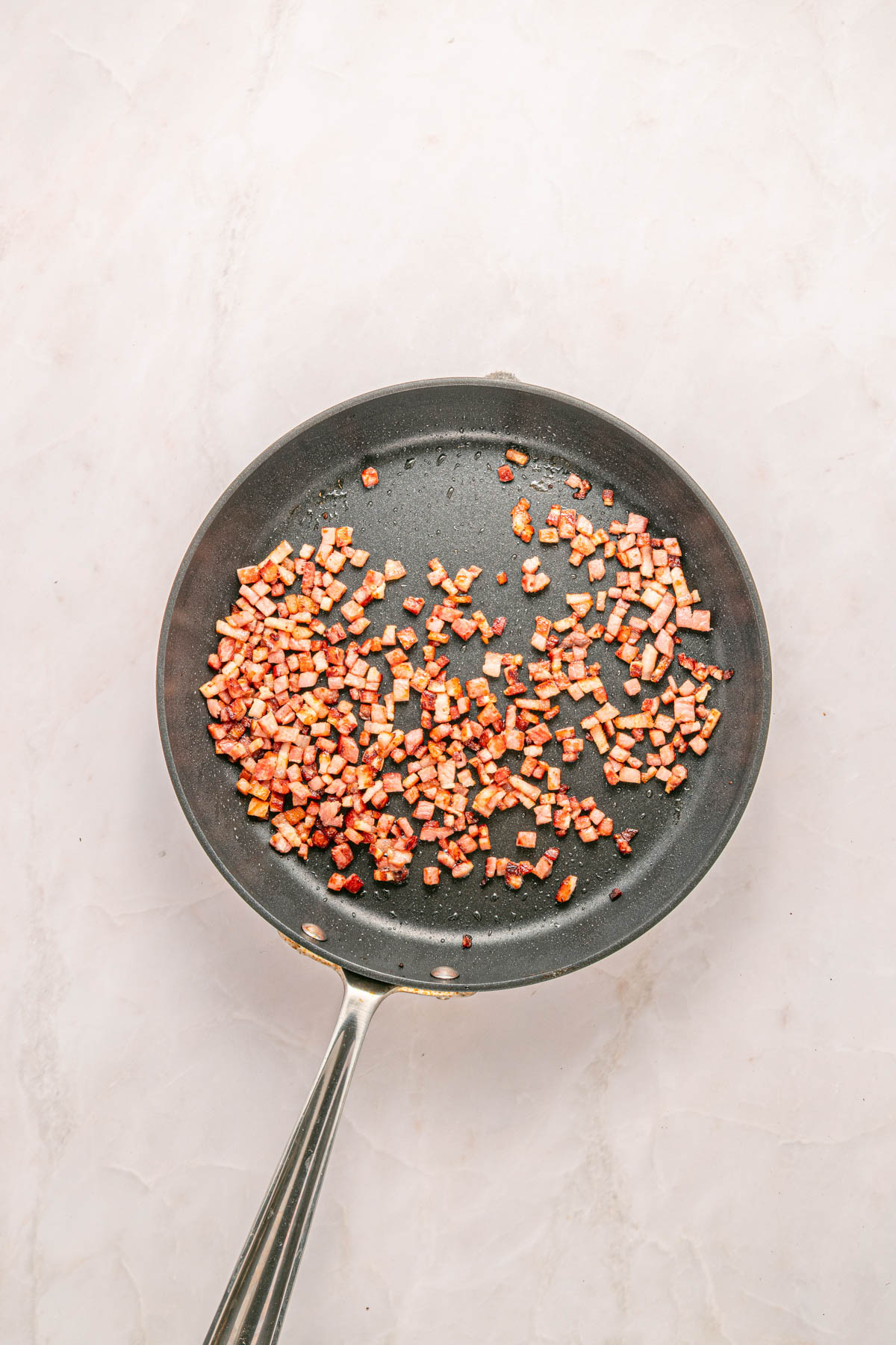 A frying pan with small diced pieces of cooked bacon spread out on its surface, placed on a light-colored countertop.
