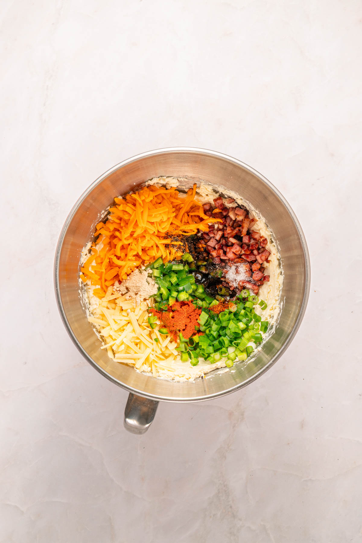 A metal mixing bowl containing shredded cheddar and mozzarella cheese, chopped green onions, diced bacon, flour, and spices on a light countertop.