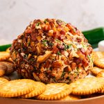 A cheese ball covered in chopped nuts and herbs is surrounded by round crackers on a platter.