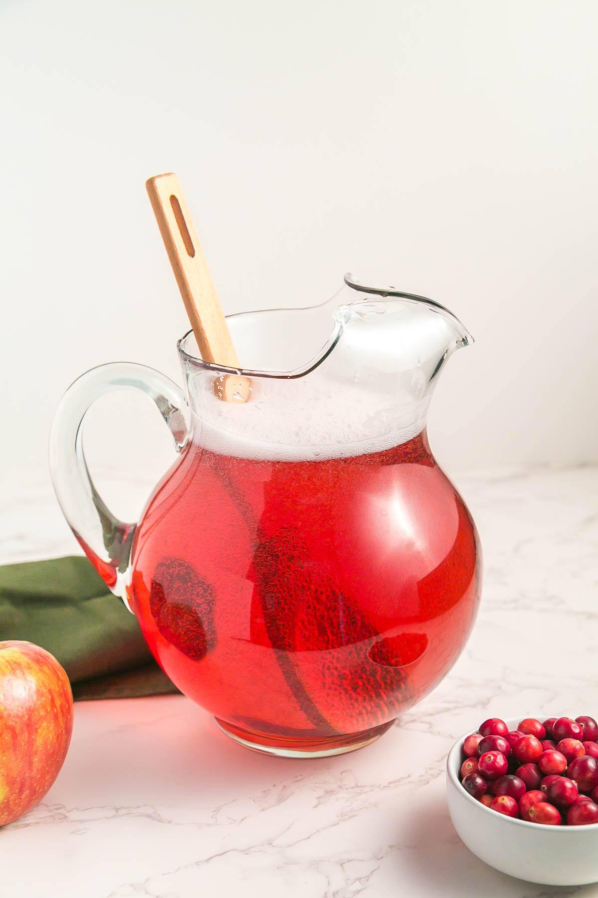 A glass pitcher filled with red juice and a wooden spoon, next to an apple, a green cloth, and a small bowl of cranberries on a marble surface.