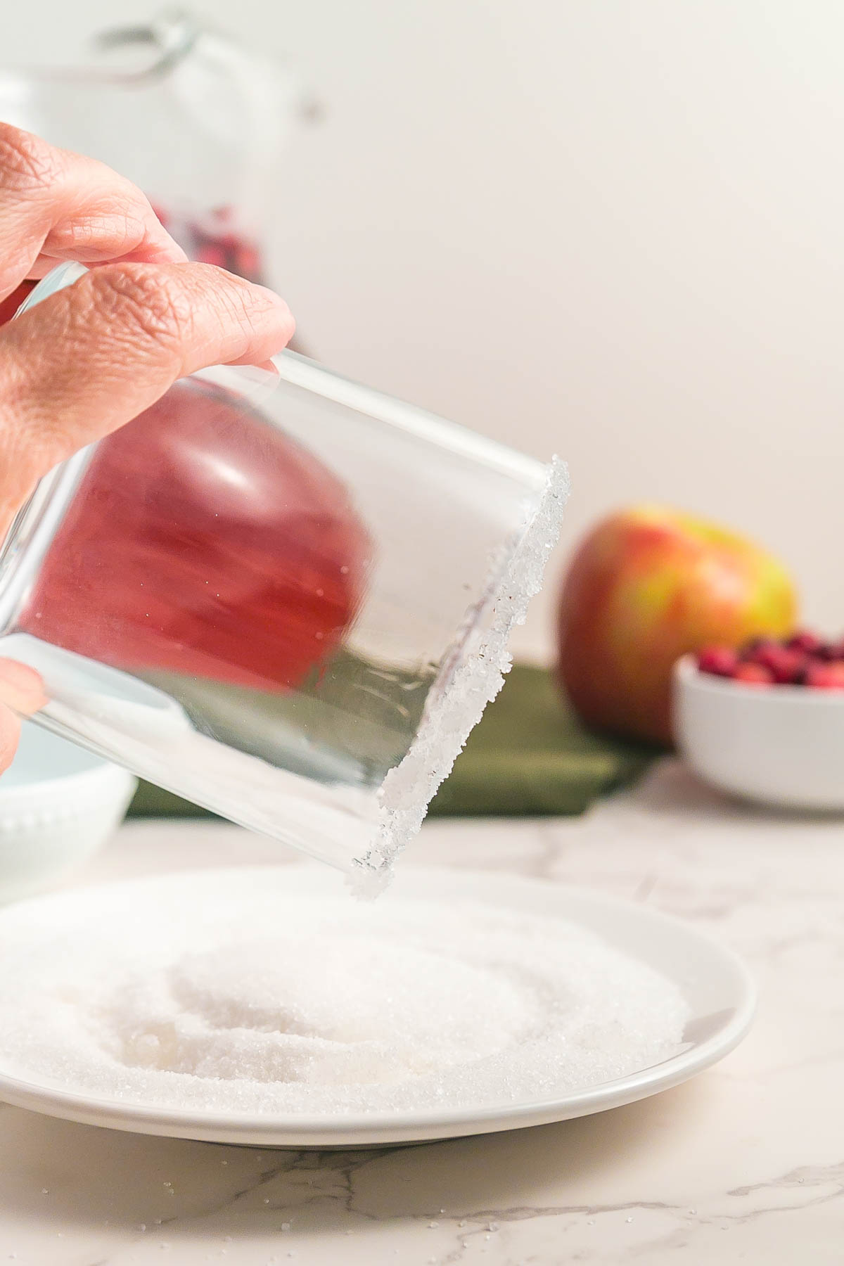 A hand dips the rim of a glass into a plate of sugar, with fruit and a bowl of cranberries in the background on a marble surface.