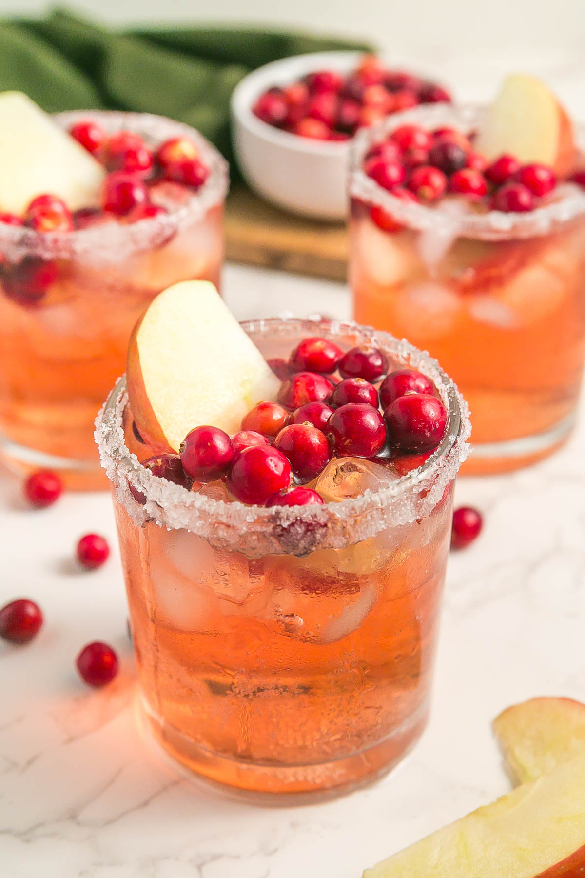 Three glasses of iced cranberry cocktails with sugared rims, garnished with fresh cranberries and apple slices, set on a white surface with extra cranberries and a green cloth in the background.