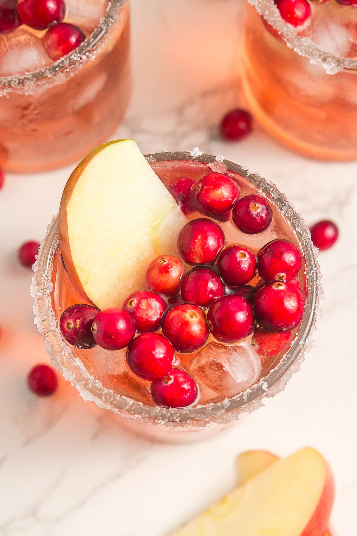A glass of iced pink beverage garnished with cranberries and a slice of apple, with a sugared rim, viewed from above. Other glasses and cranberries are visible in the background.