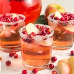 Three glasses of iced punch with sugared rims, garnished with apple slices and cranberries, are surrounded by scattered cranberries and apple pieces on a white surface.